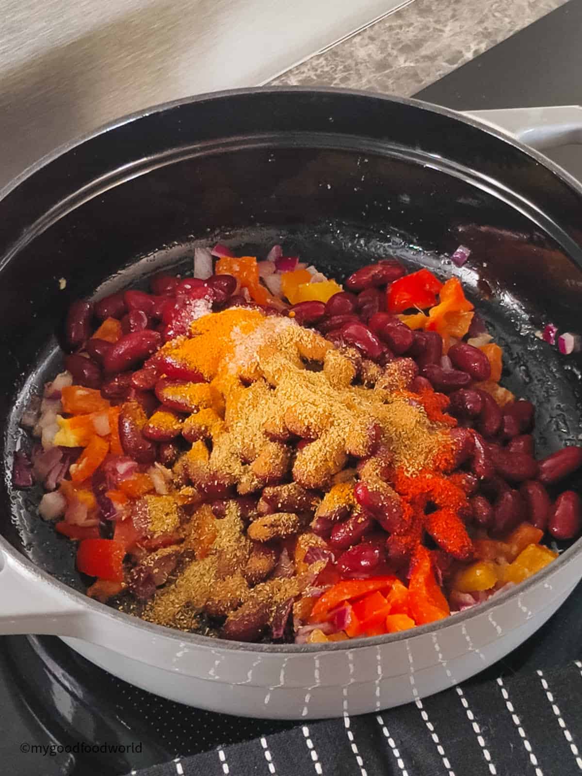 Kidney beans, peppers, and onion are topped with turmeric, cumin, and paprika before cooking in a black pan.