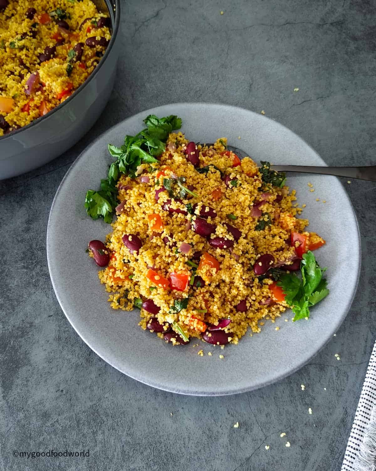 Spicy couscous recipe with kidney beans, peppers, and cilantro served on a grey plate, with the pan of salad in the background.