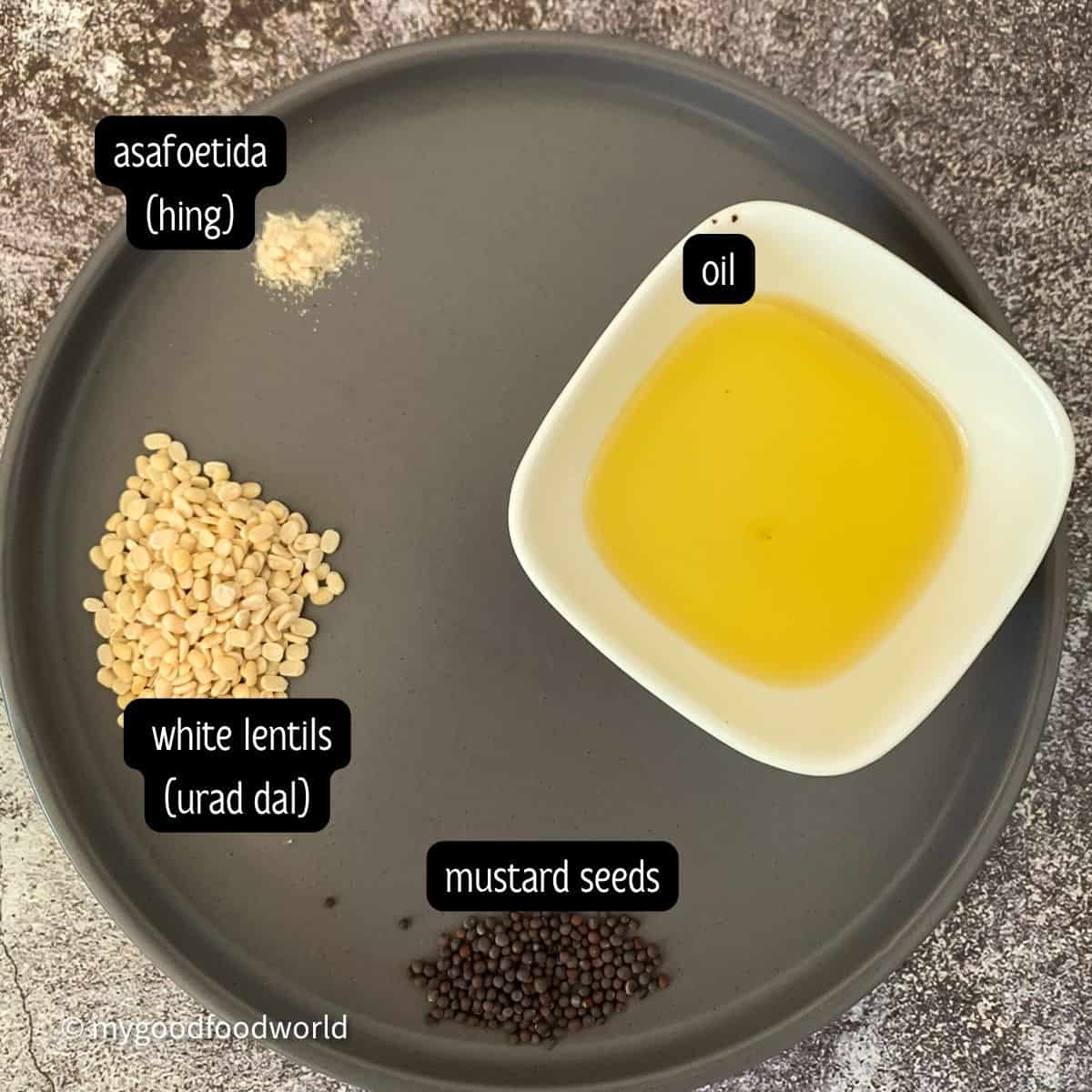 Tempering ingredients for dosakaya pachadi - urad dal, mustard seeds, oil, and asafoetida powder in a round grey plate.