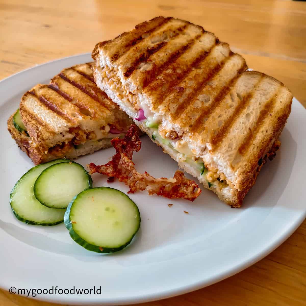 Paneer cutlet toastie toasted in a sandwich press, cut in half on a white plate, with melted cheese, cucumber, and red onion.